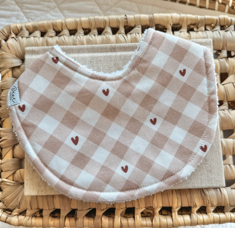 Handmade baby bib featuring a beige and white gingham print with scattered tiny brown hearts, styled flat in a woven basket with a neutral linen backdrop.