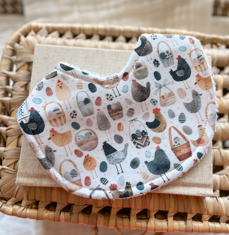 Handmade baby bib with illustrated chickens, farm eggs, and baskets in soft earthy tones, backed with absorbent terry towelling; perfect for teething or newborn messes.