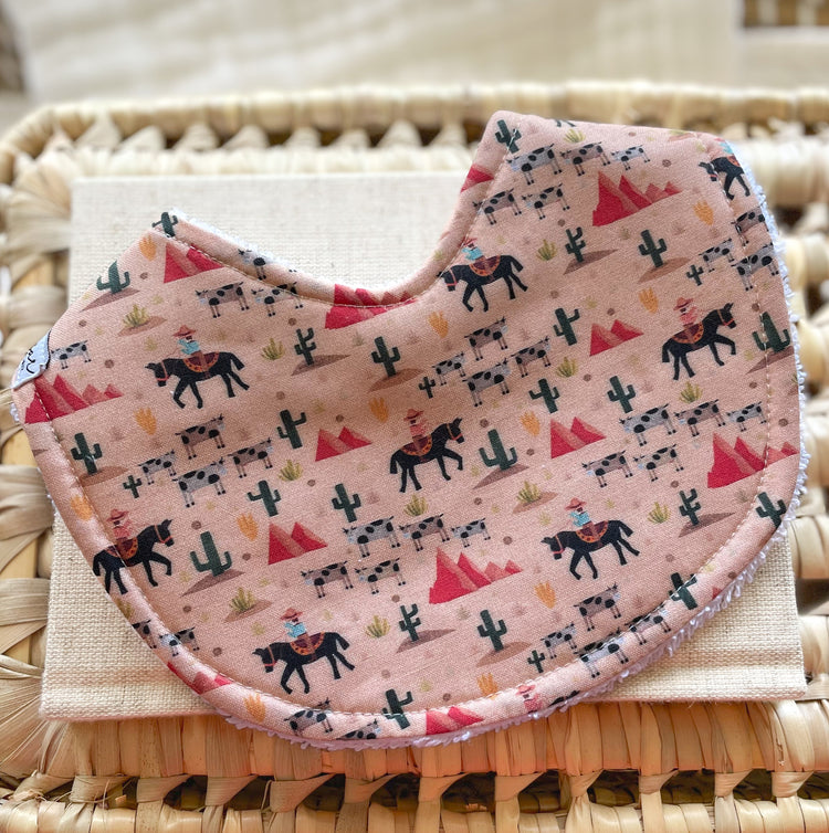 Handmade baby bib with cowboys riding horses, black and white cows, cacti, and desert hills on a dusty pink background; absorbent terry towelling backing, ideal for newborns and teething babies.