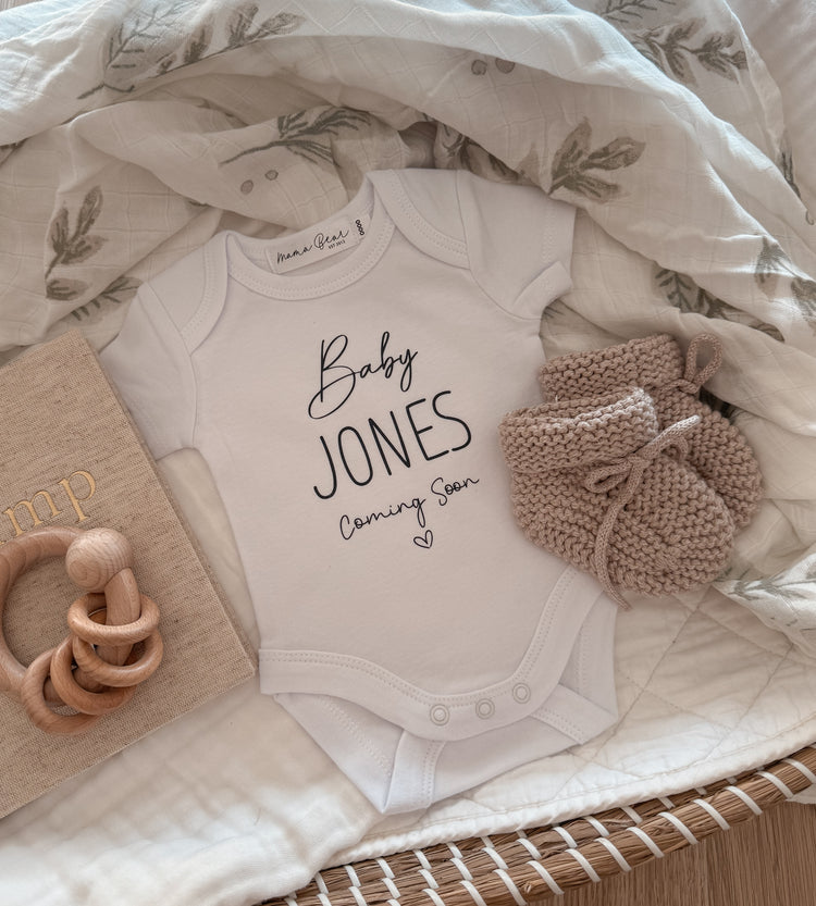 Neutral flat lay of a white baby onesie with ‘Baby JONES Coming Soon’ in a mix of script and capital font. Styled in a wicker basket with a beige floral blanket, knitted baby booties, and a ‘bump’ pregnancy journal. Perfect for a pregnancy reveal or flat lay announcement.