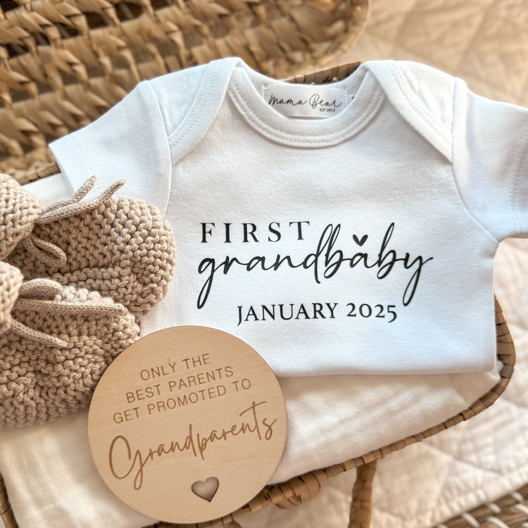 Flat lay of a personalised white baby onesie featuring the phrase “First Grandbaby January 2025” in a mix of serif and script fonts with a small heart underneath. The onesie is placed in a wicker basket with a soft cream baby wrap, beige knitted booties, and a wooden disc engraved with “Only the best parents get promoted to grandparents.” A sentimental and elegant pregnancy reveal gift for first-time grandparents.