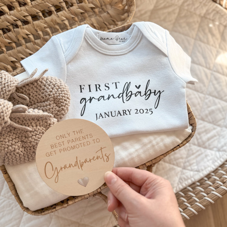 Flat lay of a white baby onesie printed with “First Grandbaby January 2025” in black text, styled inside a wicker basket on a soft cream blanket. The setup includes a round wooden plaque that reads “Only the best parents get promoted to grandparents” and a pair of beige knitted booties. A thoughtful and neutral-toned pregnancy announcement idea for first-time grandparents.