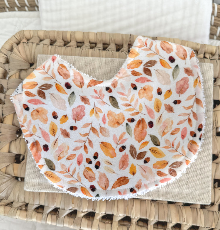 Contoured baby bib with a warm autumn leaf print in golden yellow, rust, olive green, and blush pink on white cotton, designed for newborns and handmade with soft absorbent fabric.