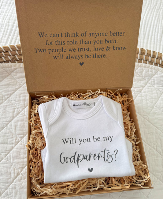 A kraft brown gift box with 'We can't think of anyone better for this role than you both. Two people we trust, love & know will always be there..." on the lid and inside a onesie that says 'Will you be my Godparents?'