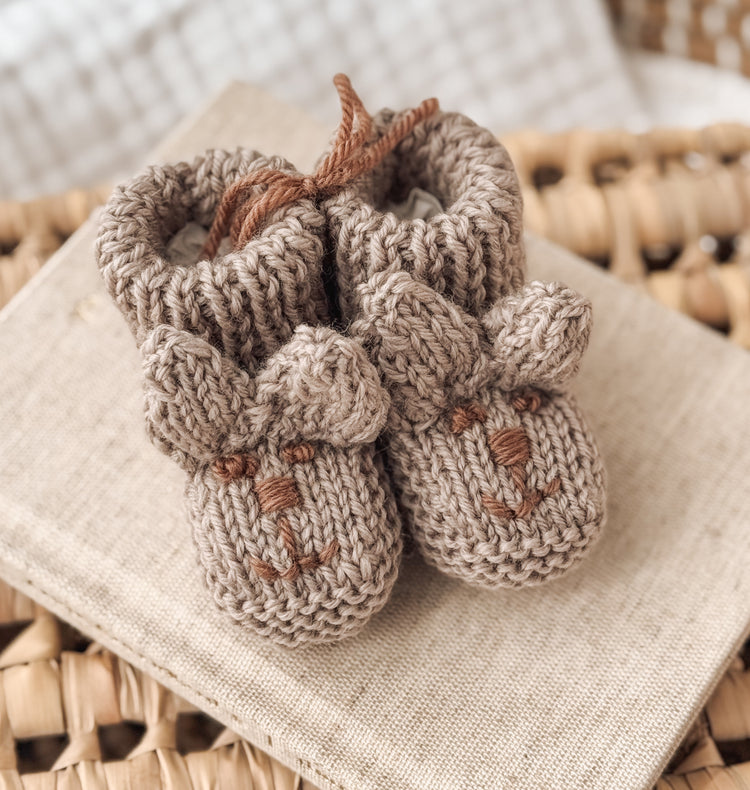 A pair of handmade knitted baby bunny booties. Embroided a little brown face. The booties are a soft brown, beige colour with little bunny ears. Perfect gift for a baby shower or newborn.