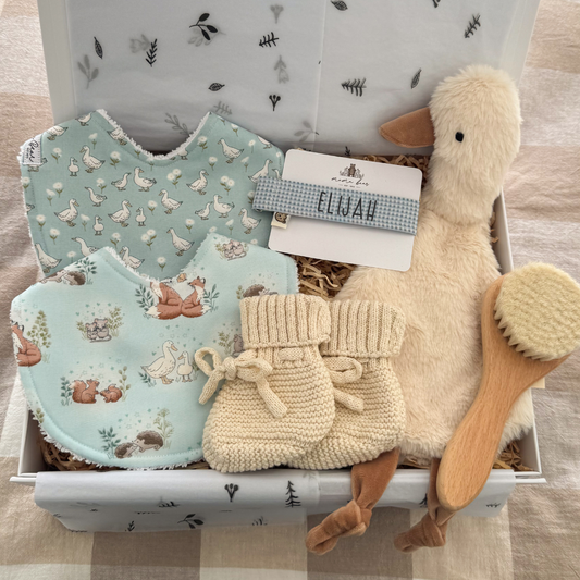 A beautifully curated baby gift box open to reveal two handmade sage mint fabric bibs with woodland and duck prints, a pair of tan leather baby moccasin sandals, a fluffy cream plush goose soft toy, and a blue gingham personalised dummy clip, all nestled in a white keepsake gift box with straw filler and a gold “BABY” gift tag