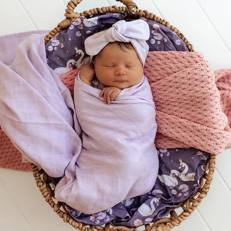 Newborn baby swaddled in a purple blanket with a floral headband, lying in a woven basket with pink and purple blankets.