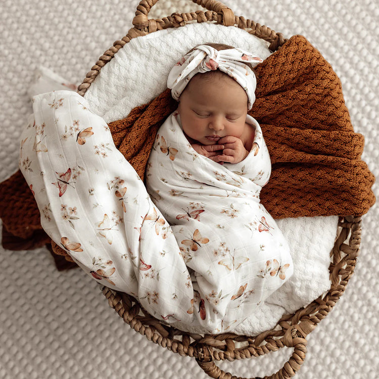 Newborn baby girl wrapped in a floral butterfly blanket with a brown knitted wrap, lying in a wicker basket on a textured surface.