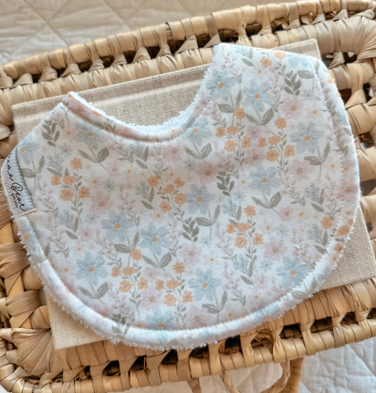 Flat lay of a handmade baby bib with a soft floral print in pastel shades of peach, pink, and green. The bib is styled on a woven tray with a neutral linen background.