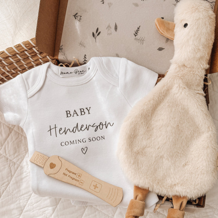 Custom baby announcement gift box with “Baby Henderson Coming Soon” onesie, soft duck comforter, and wooden pregnancy test – a special pregnancy reveal idea for gifting in person or mailing to family.