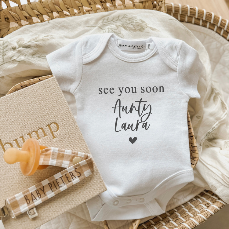 Flat lay of a white baby onesie with the message “See you soon Aunty Laura” in a mix of serif and handwritten script fonts, printed in black with a heart underneath. Styled in a wicker basket with a leafy muslin wrap, beige “bump” journal, a gingham dummy clip personalised with “BABY PIETERS,” and a caramel dummy. A personalised pregnancy announcement gift for aunties or loved ones—custom names available for any recipient.