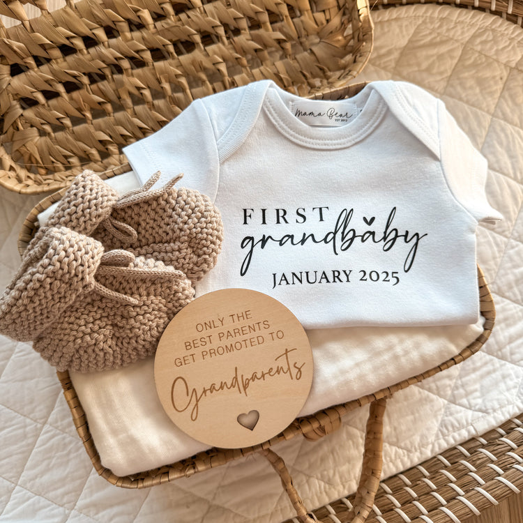 Detailed flat lay of a personalised baby announcement onesie that says “First Grandbaby January 2025.” The onesie is displayed in a wicker basket alongside soft beige baby booties and a wooden sign for grandparents. A simple and heartwarming baby reveal idea for social media or surprise gift boxes.