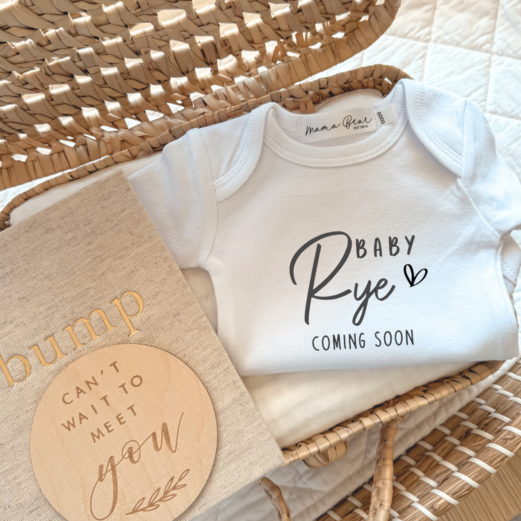 Flat lay of a white personalised baby onesie with the text “Baby Rye Coming Soon” in a mix of playful script and serif fonts, featuring a heart detail. The onesie is nestled in a wicker basket with a neutral “bump” pregnancy journal and a round wooden plaque that reads “Can’t wait to meet you.” A custom baby announcement outfit, perfect for social media reveals, flat lays, or gifting long-distance loved ones.
