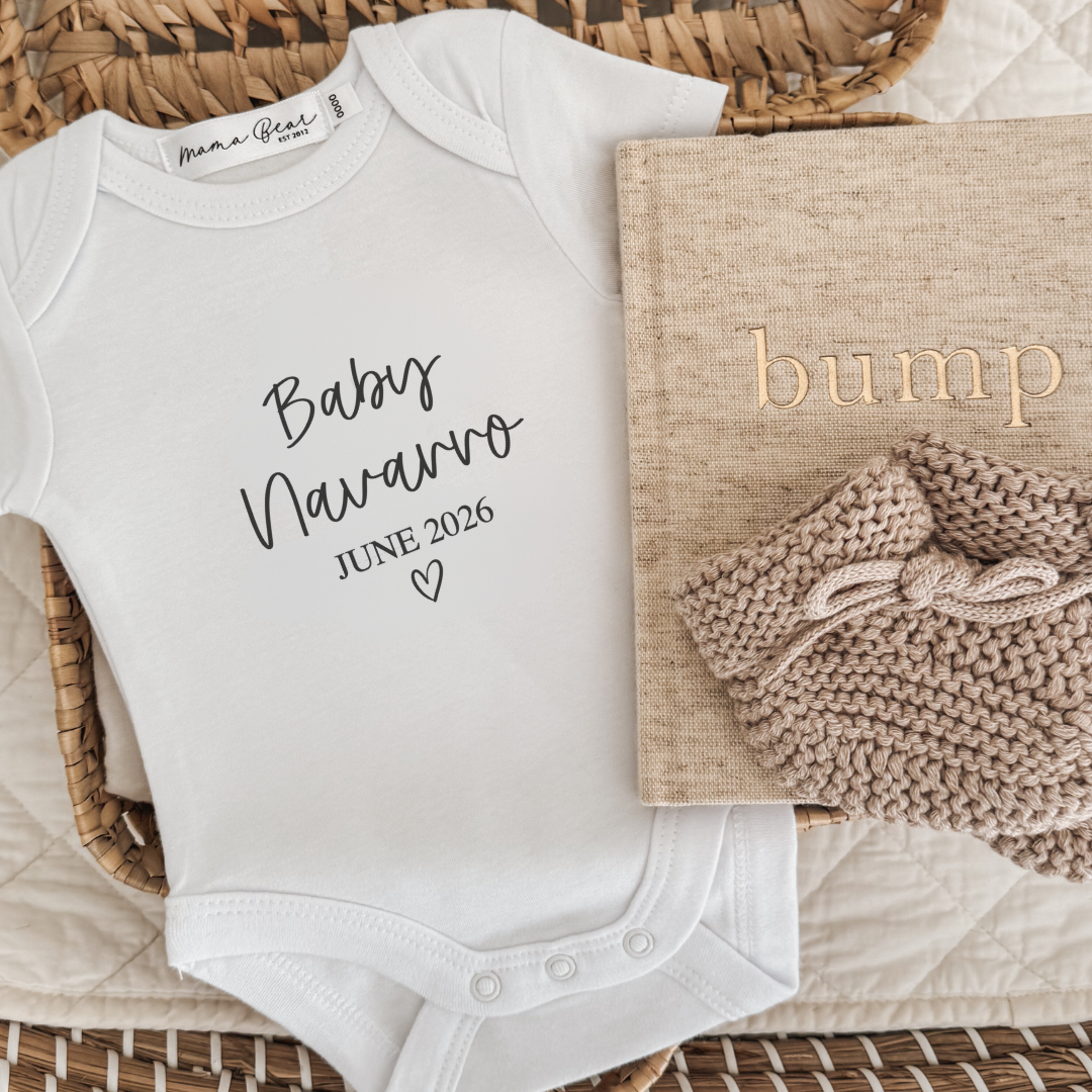 White baby onesie with personalized text, placed on a textured surface with a book and knitted item.