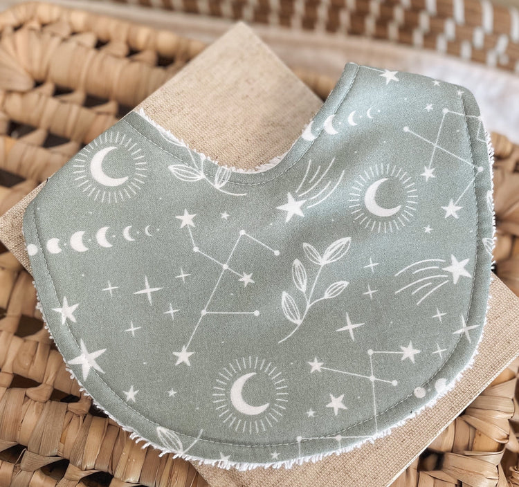 Sage green baby bib with a celestial print of moons, stars, constellations, and botanicals; handmade with soft cotton and absorbent terry towelling backing, ideal for newborns and teething.