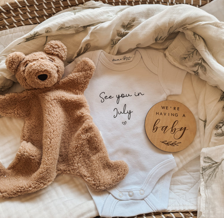Flat lay baby announcement onesie See you in July'. Pictured with a teddy bear and 'we're having a baby' wooden plaque.