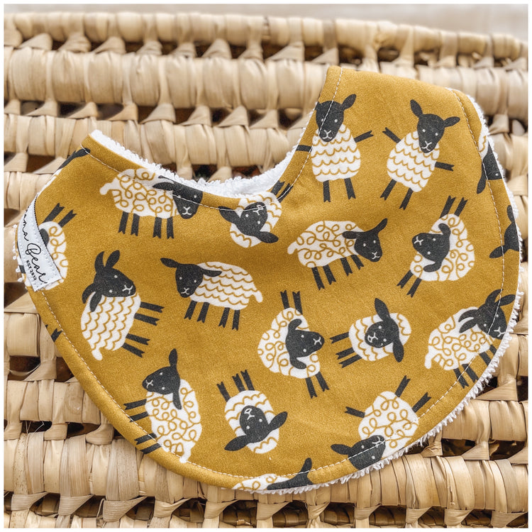 Baby bib with black and white sheep print on a mustard yellow fabric, styled on a woven basket.