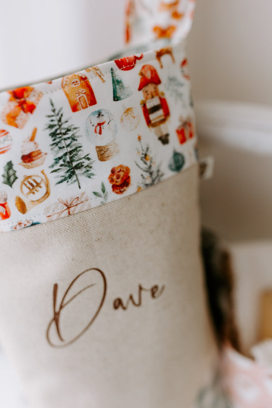 Handmade personalised Christmas stocking in natural linen with the name “Dave” in brown script font, featuring a festive Santa Claus print cuff with reindeer, Christmas trees and gifts, and finished with sage green gingham heel and toe patches. Rustic farmhouse style holiday stocking, custom made in Australia, ideal for family or children’s Christmas décor.