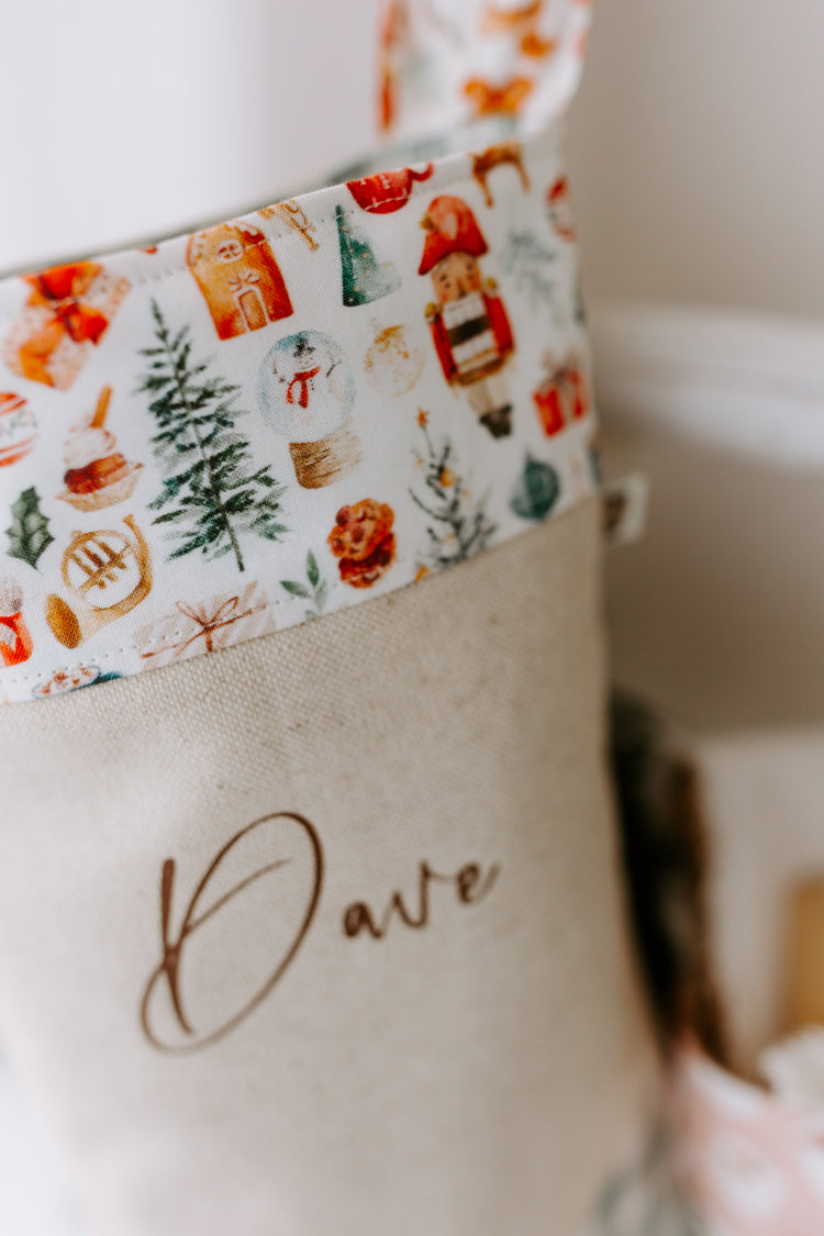 Handmade personalised Christmas stocking in natural linen with the name “Dave” in brown script font, featuring a festive Santa Claus print cuff with reindeer, Christmas trees and gifts, and finished with sage green gingham heel and toe patches. Rustic farmhouse style holiday stocking, custom made in Australia, ideal for family or children’s Christmas décor.