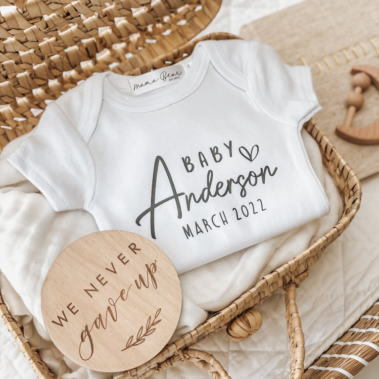 Flat lay of a white baby onesie with ‘Baby Anderson March 2022’ printed in script and serif fonts, styled in a wicker basket with a round wooden disc engraved with “We never gave up.” A meaningful and personalised baby announcement onesie, perfect for pregnancy reveals, especially after infertility or IVF journeys.