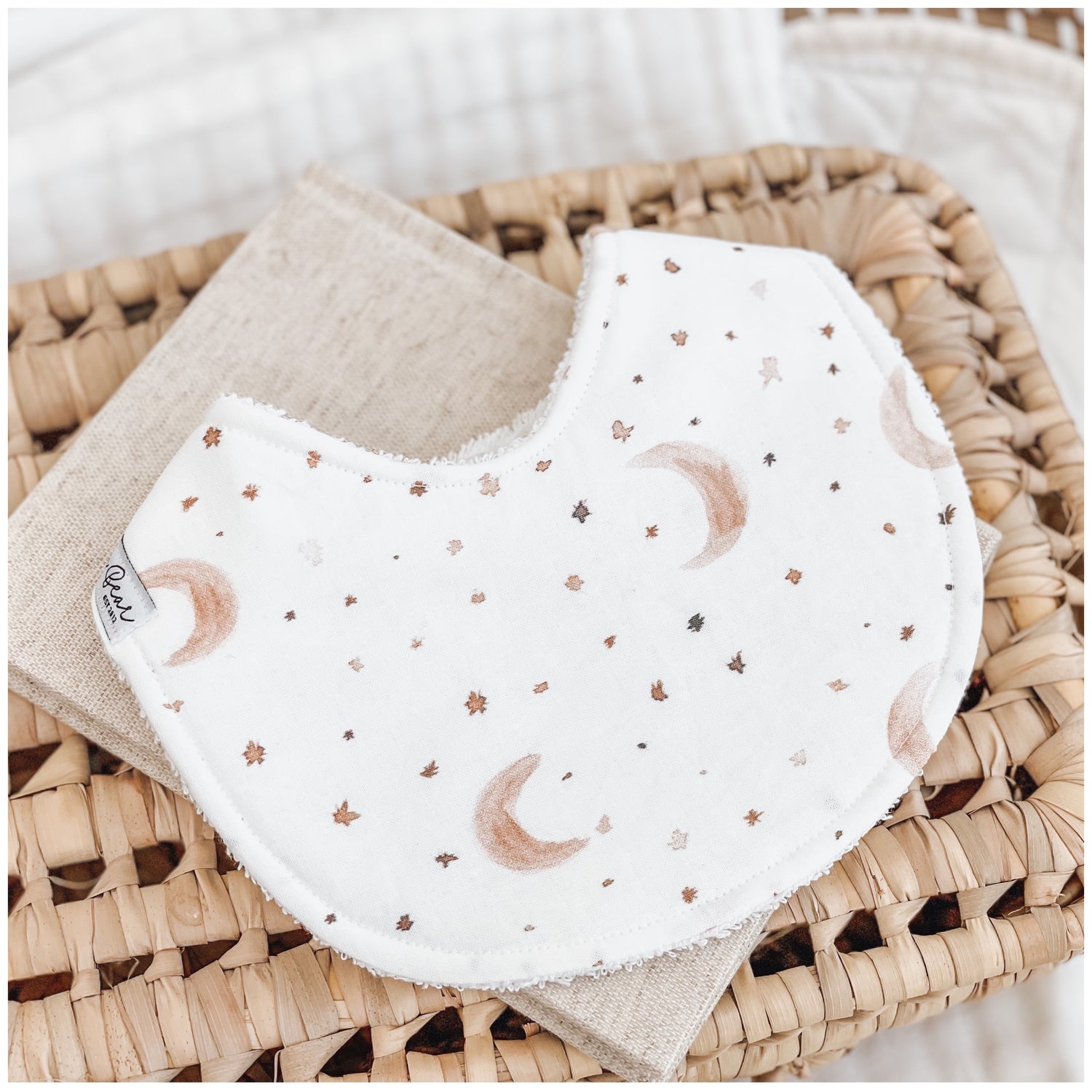 Handmade baby bib with a soft moon and star print in earthy browns and beige on a white base. Bib is backed with beige terry towelling and displayed on a woven basket.