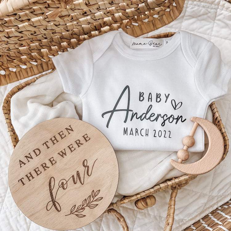 Personalised baby announcement onesie featuring the name “Baby Anderson” and “March 2022,” placed in a neutral-toned flat lay. Styled with a wooden disc reading “And then there were four” and a wooden crescent moon rattle, this onesie makes a perfect custom gift for baby reveals or social media announcements.