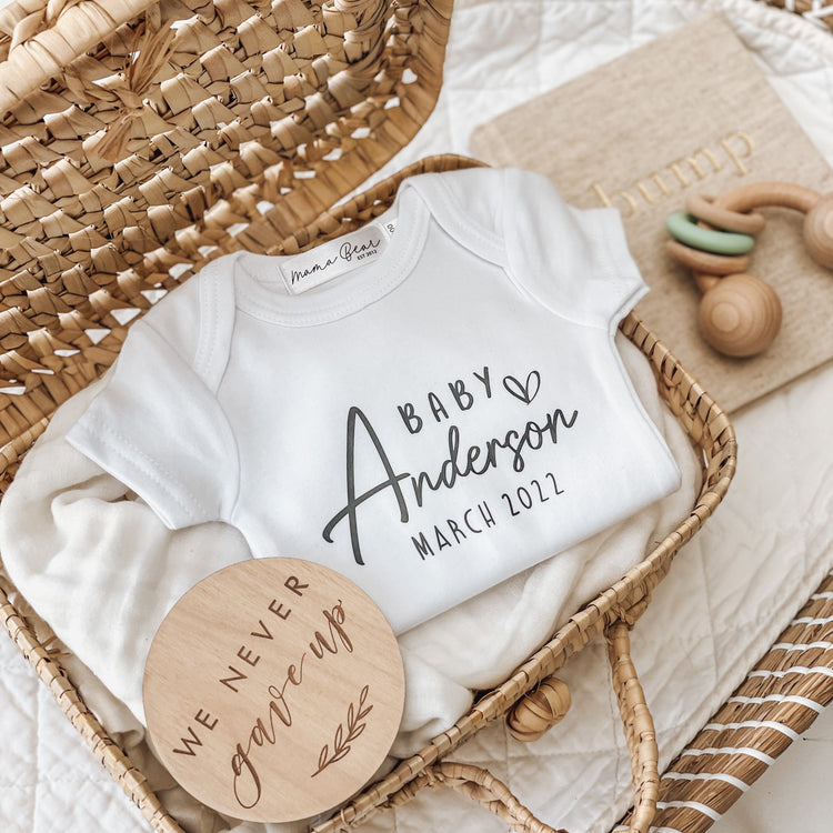 Personalised white onesie with “Baby Anderson March 2022” printed across the chest, laid flat in a woven basket with a wooden “We never gave up” disc, bump journal, and wooden toy. A beautiful baby announcement idea to mark a long-awaited pregnancy or to surprise family and friends with the news.