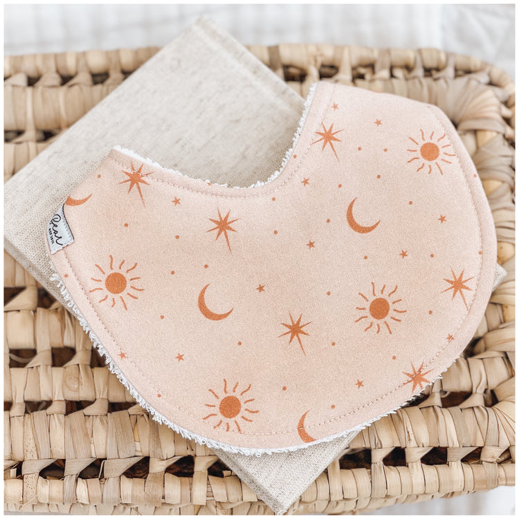 Handmade baby bib with a soft peach background and scattered burnt orange sun, moon, and star illustrations. Bib is backed with beige terry towelling and styled on a woven basket.