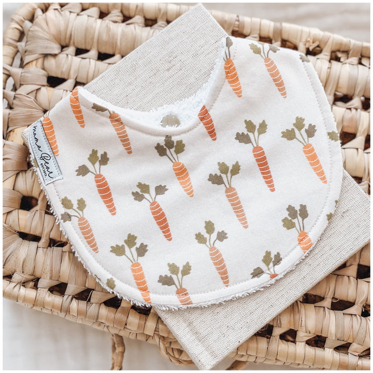 Neutral baby bib with an all-over print of orange carrots and green tops on a cream background, styled on a woven basket.