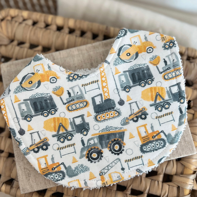 Flatlay of a handmade baby bib with a construction-themed print. The design includes yellow and grey vehicles such as diggers, dump trucks, cement mixers and cranes on a white background. The bib is cotton with a soft terry towelling back for absorbency.