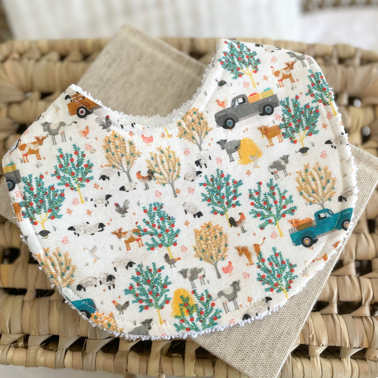 Flatlay of a handmade baby bib with a farm-themed print. The design features scattered utes, sheep, cows, trees, and chickens on a cream background. The bib is made from cotton with an absorbent white terry towelling back.