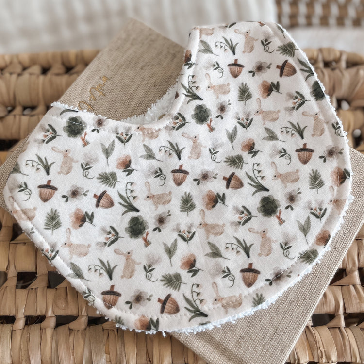 Neutral-toned baby bib with a woodland pattern of rabbits, acorns, mushrooms, leaves, and greenery on a cream background. Backed with terry towelling, ideal for dribbles, teething, and newborn feeding.