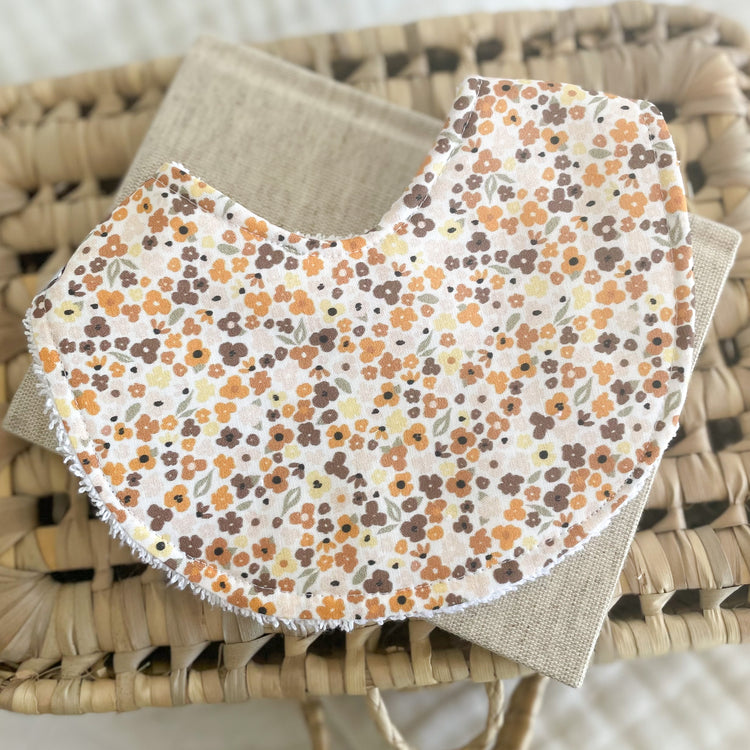 Handmade baby bib with tiny scattered flowers in brown, mustard yellow, and burnt orange on a soft cream background. Laid flat on a neutral fabric and woven basket.