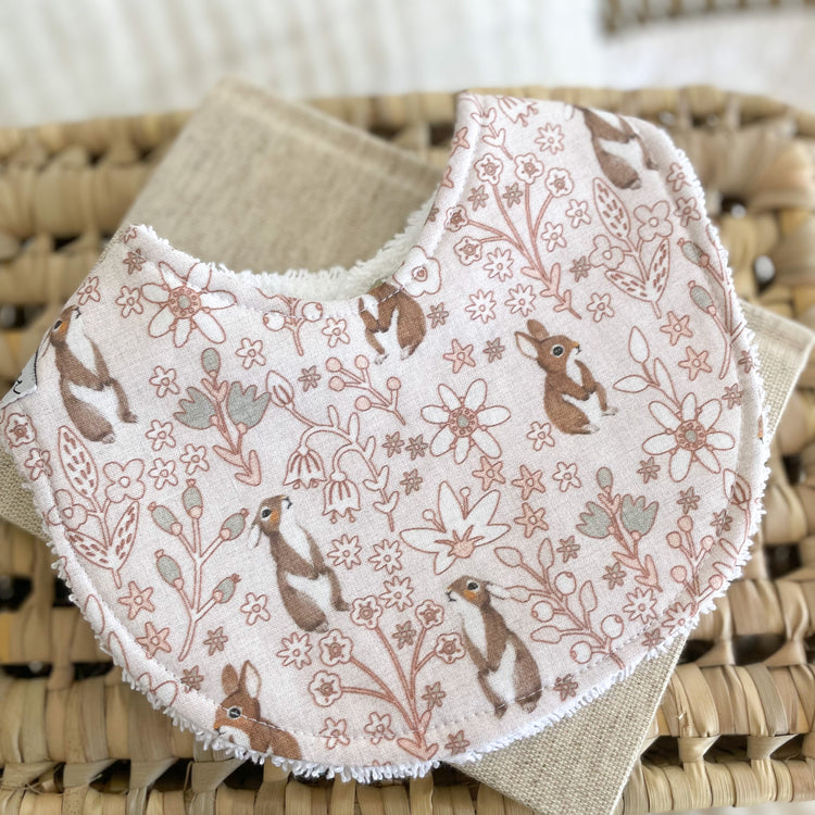 Flatlay of a handmade baby bib with a light pink background, covered in dainty floral line drawings and brown bunny rabbits. The bib is made from soft cotton with a white terry towelling back for absorbency.