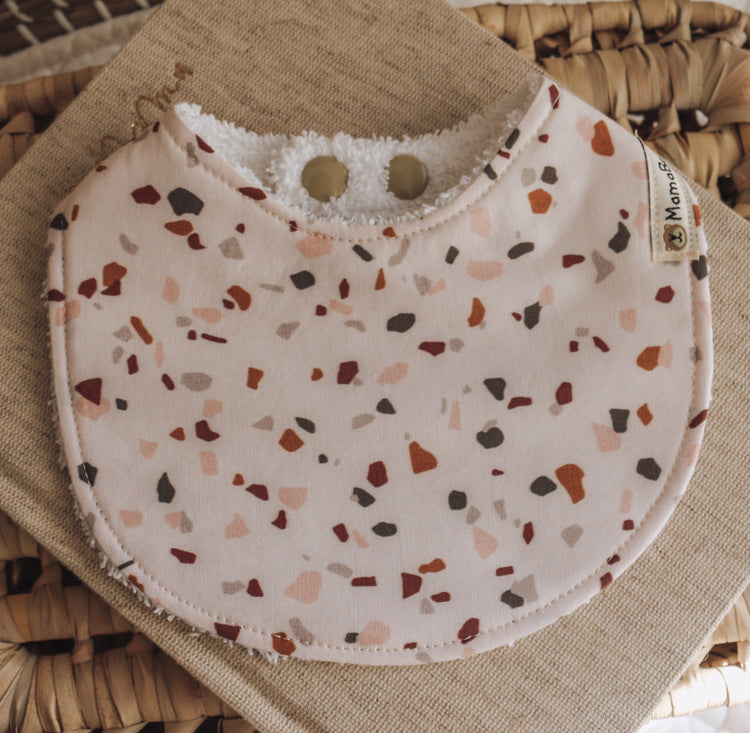 Handmade baby bib with a terrazzo-style abstract print in warm peach, brown, and clay tones on a soft blush background, styled on a natural woven backdrop.