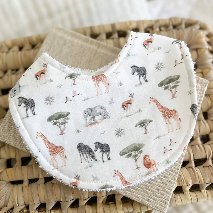Flatlay of a handmade baby bib with a safari animal print including giraffes, elephants, zebras, birds, and trees on a soft neutral background. Made with cotton and white terry towelling backing.