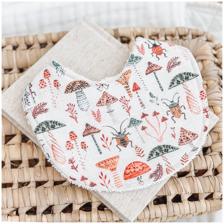 Handmade baby bib with a woodland pattern showing red, orange, and green mushrooms, forest leaves, and beetles on a soft neutral background. Bib is backed with beige terry towelling and styled on a woven basket.