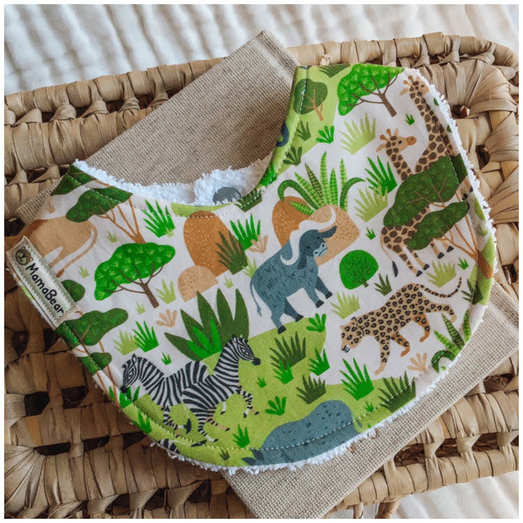 Baby bib with a bright safari print featuring jungle animals like elephants, giraffes, leopards, and zebras on a green and cream background. Displayed on a woven basket with a neutral cloth — a handmade, gender-neutral bib ideal for adventurous little ones.
