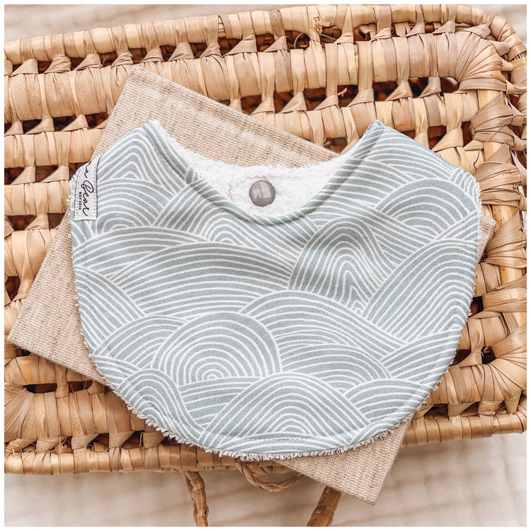 Seafoam blue baby bib featuring white curved line waves in a layered, ripple-style pattern, resting on neutral-toned fabric in a wicker basket.