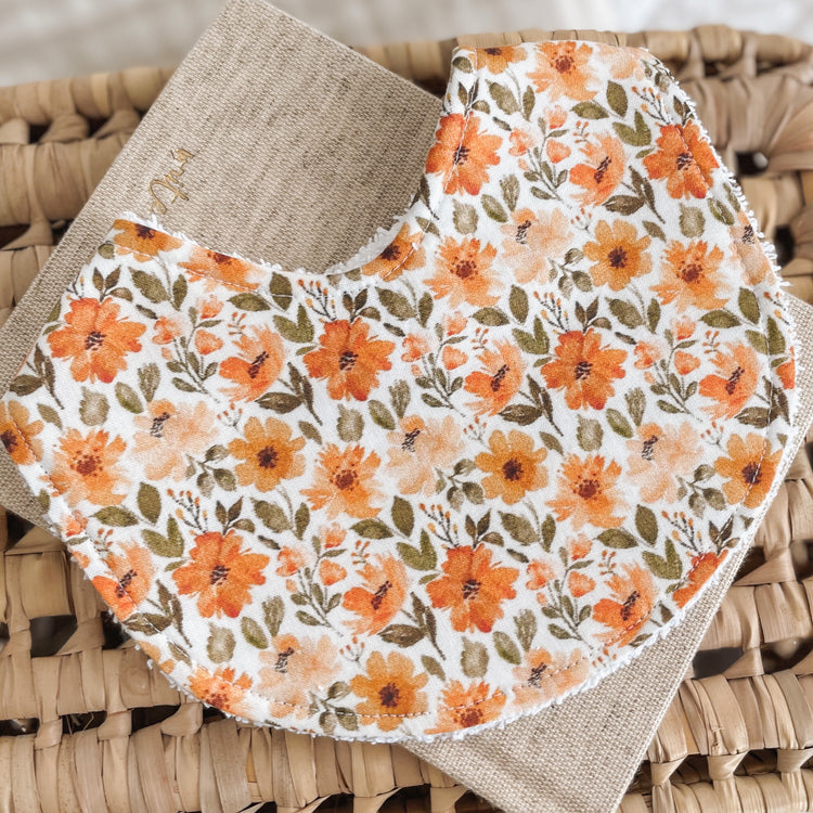 Baby bib with a bold floral print featuring burnt orange, peach, and golden yellow flowers with green leaves on a cream background. Made with absorbent terry towelling and ideal for newborn dribbles and teething.