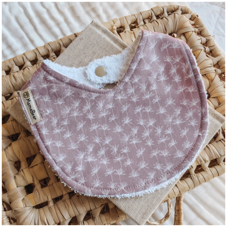 Baby girl bib in soft lilac with a fine white micro daisy print. Photographed on a woven basket with a neutral cloth — a handmade floral bib for newborn girls or baby gifts.