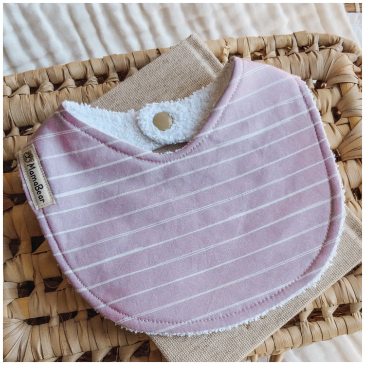 Baby bib in soft lilac with fine white horizontal stripes. Displayed on a woven basket with a natural fabric base — a handmade baby girl bib in a simple and timeless design.