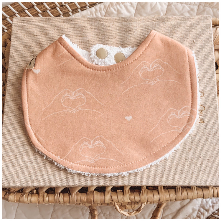Handmade baby bib in terracotta with delicate line drawings of hands forming heart shapes, backed with white terry towelling, displayed on neutral fabric with a natural woven basket background