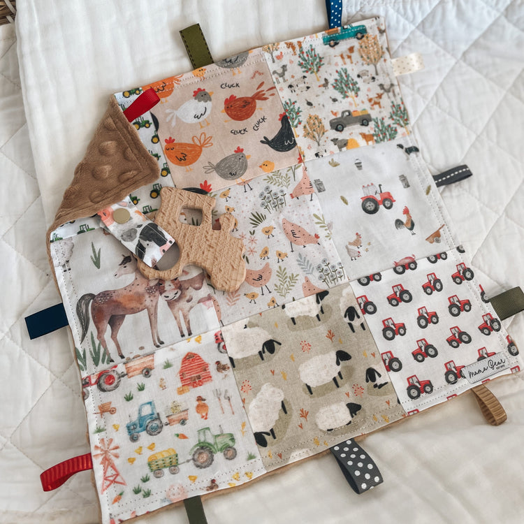 Handmade baby Taggie blankie featuring patchwork squares with farm animals, tractors, sheep, horses, chickens, and barns in warm countryside tones. Includes a wooden cow-shaped teether on a cow-print strap, tactile ribbon loops in mixed colours, and a soft minkee dot corner for sensory play. A “Mama Bear” label is stitched in the corner.