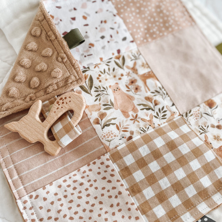 Handmade baby Taggie blanket in warm earthy tones with a mix of patchwork prints including gingham, florals, terrazzo, stripes, and woodland animals. Features a wooden deer-shaped teether attached with a matching fabric strap, textured minkee backing, and neutral-toned ribbon loops for sensory play.
