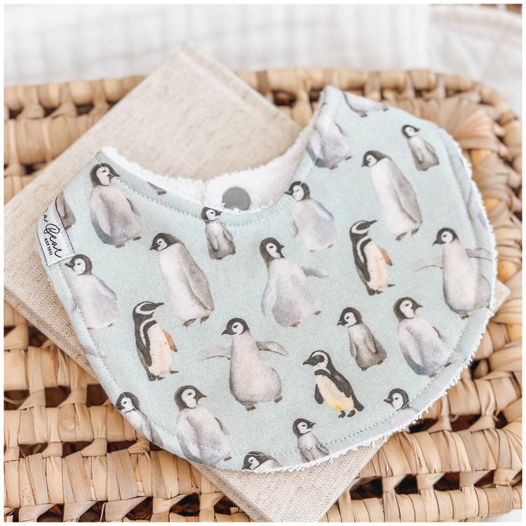 Handmade baby bib with a cute penguin pattern showing adult and baby penguins on a light blue background. Bib is backed with beige terry towelling and displayed on a woven basket.