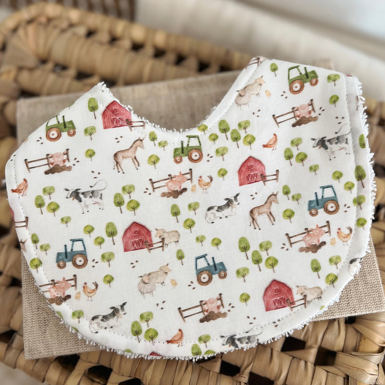 Flatlay of a handmade baby bib with a farm-themed pattern. The design includes cows, sheep, horse, chickens, blue and green tractors, red barns, and small trees on a light cream base. The bib has a cotton front and a soft terry towelling back for absorbency.
