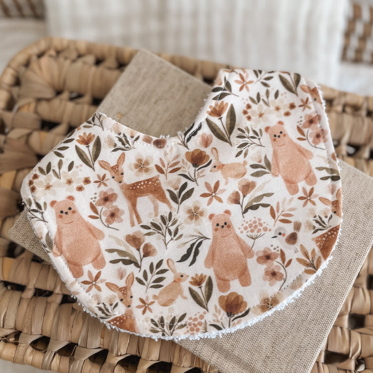 Neutral baby bib with a woodland animal pattern including bears, deer, bunnies, and florals in warm tan, brown, and rust tones on a light background. Absorbent terry backing ideal for newborn messes and teething.