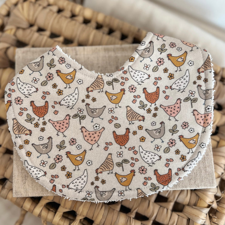 Handmade baby bib with illustrated chickens and scattered florals in earthy tones on a neutral background; backed with absorbent terry towelling, ideal for newborns and everyday use.