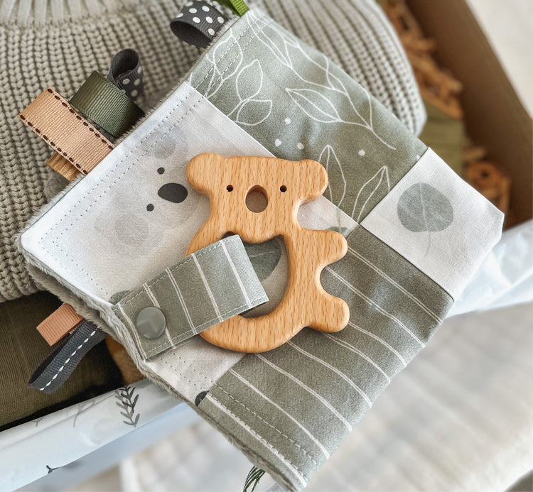 Close-up of a sage green baby Taggie blankie featuring Australian koala and eucalyptus leaf prints, stripe and leaf patterns, and a wooden koala-shaped teether attached with a matching fabric strap. Surrounded by textured ribbon loops in earthy tones.